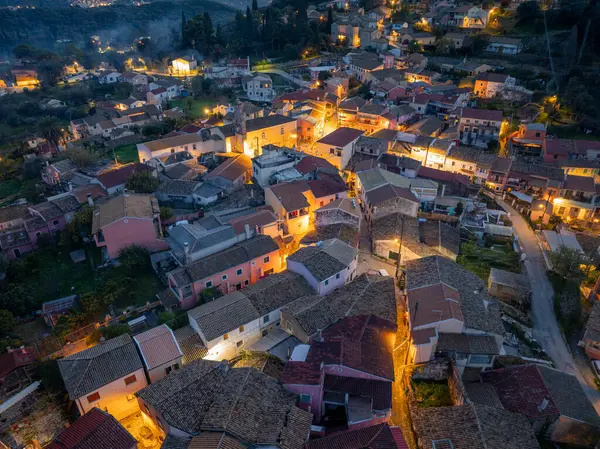 Yuvarlanan tepelerin arasına kurulmuş bir köyün yukarıdan görünüşü. Çatı kiremitleri sokak lambalarıyla aydınlatılmış binalar.