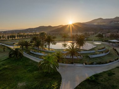 Huzurlu bir manzara, güneşin bir dağ sırasının üzerinden doğuşunu gösterir. Sabah ışığı altında, yemyeşil palmiye ağaçlarıyla çevrili bir gölette yansıtır..