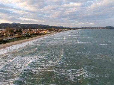 Yukarıdan manzaralı bir plaj, sakin bir sörf sahili, kıyı boyunca uzanan antika binalar ve arka planda dağlar..