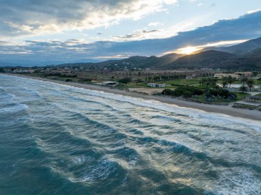 Güneşin ufukta battığı, yemyeşil tepeler ve yemyeşil kumsallarla dolu, dalgalı bir kumsalla buluşan dalgaların olduğu sakin bir sahil sahnesi..