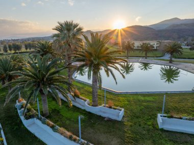 Palmiye ağaçlarının bulunduğu sakin bir manzaranın havadan görünüşü, günbatımında uzak dağlara bakan ağaçları yansıtan sakin bir havuz..