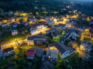 Yüksek açılı bir gece manzarası, evlerden ve sokak lambalarından yayılan sıcak ışıklarla parıldayan, yemyeşil tepelerin arasında yuva yapmış tuhaf bir köyü gözler önüne seriyor..