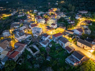 Yüksek açılı bir manzara, Alacakaranlık 'ta ilginç bir köy manzarası yakalıyor, kırmızı kiremitli çatıları ve yeşil bir manzarada parlayan sokak lambaları olan evleri gösteriyor..