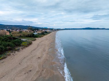 Sakin sahil manzarası, kumlu sahil dalgaları ve bulutlu bir gökyüzünün altında uzak bir kasaba..
