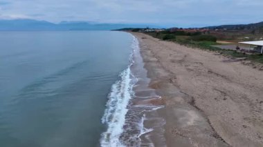 Sakin bir kumlu sahil manzarası sakin mavi sularla buluşuyor, yeşil kıyı bitki örtüsü ve uzak tepeler.