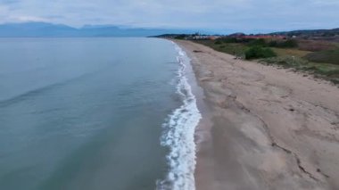 Sakin bir kumlu sahil manzarası sakin mavi sularla buluşuyor, yeşil kıyı bitki örtüsü ve uzak tepeler.