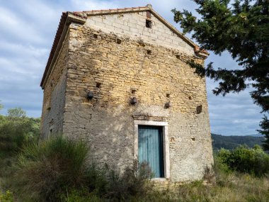 Çarpıcı mavi kapısı olan yıpranmış bir taş yapı, parçalı bulutlu bir gökyüzünün altında, yuvarlanan tepelerin zeminiyle çerçevelenmiş uzun otların arasında duruyor..