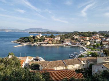 Bu görüntü, sakin bir körfezi çevreleyen büyüleyici bir kıyı kasabasını gösteriyor, limanlı tekneler ve mavi gökyüzünün altındaki uzak dağlardan oluşan bir zemin..