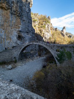 Sarmaşıklarla süslenmiş taş bir köprü, engebeli kayalıklarla çevrili taşlı bir nehir yatağının üzerinde kavisler ve parlak bir gökyüzünün altında seyrek ağaçlar. 