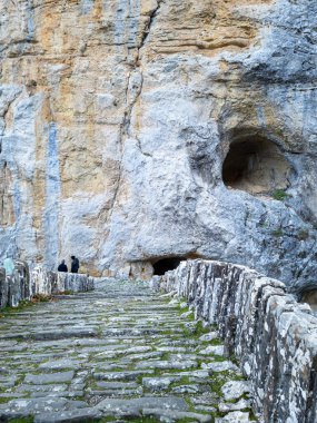 Antik bir taş yol, uzun ve katmanlı bir kaya oluşumuna doğru uzanıyor. Görünür mağaralar ve küçük insan figürleri ölçekli..