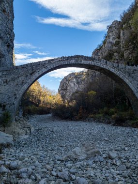 Kuru bir nehir yatağının üzerinde zarif kemerler olan eski bir taş köprü kayalarla dolu, yüksek kayalıklarla çevrili ve mavi gökyüzünün altında seyrek sonbahar yaprakları..