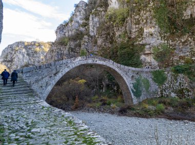 Yaş belirtileri gösteren ve sarmaşık parçalarıyla kaplı eski bir taş köprü, kayalık araziyle çevrili kuru bir nehir yatağının üzerine uzanır..