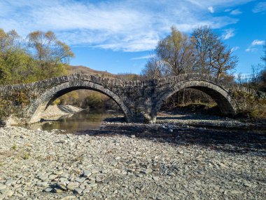 Manzaralı bir manzara, kayalık bir nehir yatağından geçen iki kemerli eski bir taş köprüyü, ağaçlarla çevrili ve canlı mavi bir gökyüzünü içerir..