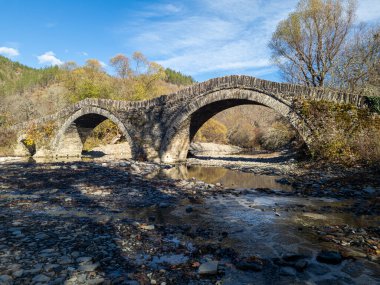 Kayalarla dolu sığ bir nehir yatağını geçen iki kemerli taştan bir köprü, sonbahar ağaçlarının ve mavi gökyüzünün zeminine kurulmuş..