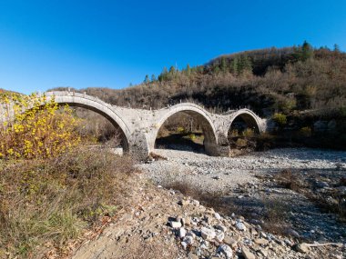 Birkaç kemerli eski bir taş köprü kayalar ve çakıllarla dolu kurumuş bir nehir yatağını geçer. Köprünün çevresi ağaçlarla çevrili..