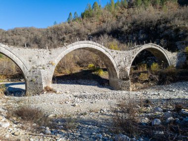 Eski, kemerli bir taş köprü taşlarla dolu kurumuş bir nehir yatağını geçer. Ağaçlarla kaplı tepeler, parlak mavi gökyüzünün altında yükseliyor..