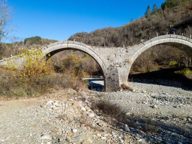 Güneşli bir günde, açık mavi bir gökyüzünün altındaki ağaçlık tepelere karşı kayalık bir nehir yatağını geçen tarihi çift kemerli taş bir köprü..