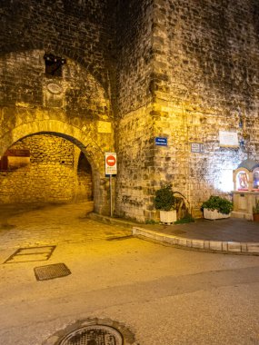 Geceleri aydınlatılmış taş bir kemer eski bir binanın köşesini aydınlatıyor, yanında süslü bir tapınak ve uyarı işareti var. Çakıl taşı.