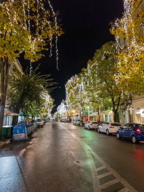 Gece bir şehir caddesi, ağaçlardaki dekoratif ışıklarla aydınlatılmış, neşeli bir ortam yaratıyor. Arabalar sokağın kenarına park edilmiş..