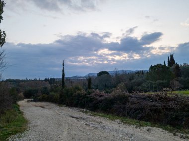 Kırsal bir yol, ufuktaki uzak dağlara doğru uzanan bulutlu bir gökyüzünün altındaki ağaçlarla birlikte bir manzara boyunca kıvrılır. Doğa görünümü.