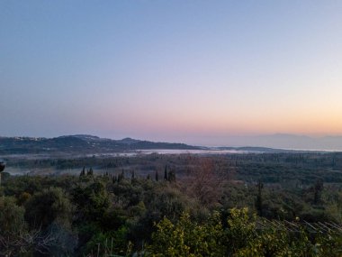 Bu sahne, vadilerde, ağaçlarda ve tepelerde, sakin ve doğal bir ortamda, pastel bir gündoğumu gökyüzünün altında gizlenmiş bir manzarayı gösteriyor..