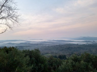 Manzaralı bir manzara, yumuşak bir sisle kaplanmış bir vadiyi gözler önüne seriyor, uzakta dağ siluetleri, hepsi şafak vakti hassas bir pastel gökyüzünün altında..