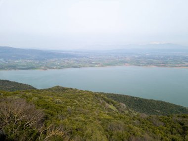 Hava görüntüsü, bulutlu bir gökyüzünün altında yeşil tepeler ve tarlalarla çevrili geniş bir gölü ve uzak dağların manzaraya derinlik kattığını gösteriyor..