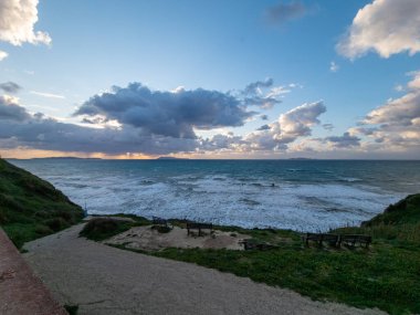 Dinamik bulutlu bir gökyüzünün altında dalgalı bir deniz manzaralı kıyı manzarası. Bir doğa yolu kompozisyona derinlik ekler.