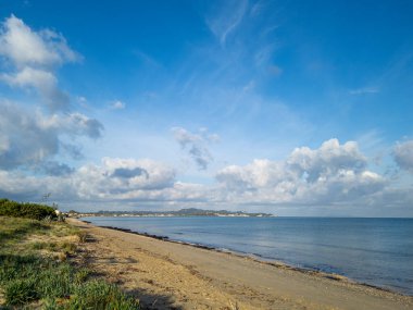 Sakin bir sahil manzarası, yumuşak dalgalar kumlu kıyılara, uzak tepelere ve güzel mavi bulutlu gökyüzüne çarpıyor..