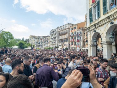 Eski binalarla çevrili bir şehir meydanını geniş bir insan topluluğu dolduruyor. Sahne kalabalık bir açık hava olayını yakalıyor..