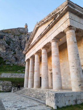 Bu resim, üzerinde deniz feneri olan bir tepenin önündeki birçok belirgin sütunu olan klasik bir binayı gösteriyor..
