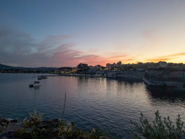 Limanda yelkenlilerin olduğu sahil manzarası, alacakaranlıkta aydınlatılmış binalar ve renkli gökyüzünün büyüleyici bir karışımı..