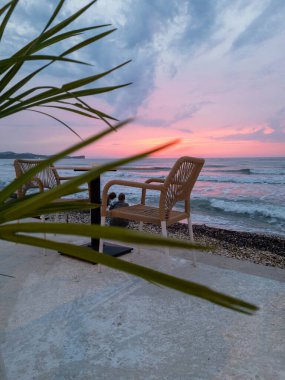 Huzurlu bir sahil sahnesinde, okyanus dalgaları üzerinde canlı bir günbatımı ile palmiye yapraklarıyla çerçevelenmiş iki dokuma sandalye ve bir masa bulunur..