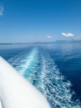 A scenic view from a moving boat, capturing the serene blue sea, distant coastline, and the boat's foamy wake under a clear sky.