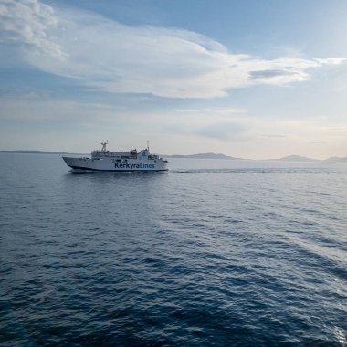 Bir yolcu feribotu, arka planda uzak tepeler olan parçalı bulutlu bir gökyüzünün altında koyu mavi bir su üzerinde hareket eder..