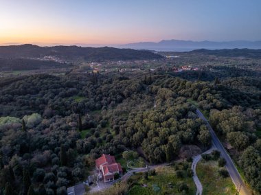 Sakin bir manzara, arka planda alacakaranlığın yumuşak renkleri ve uzak tepeleri olan sık ağaçlarla çevrili bir evi gözler önüne seriyor..