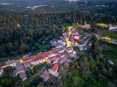 Küçük bir köyün hava görüntüsü. Binalar sık ağaçların arasında yer almaktadır. Köy, sokak ışıklarıyla aydınlatılmış kırmızı çatılı evlere sahiptir..