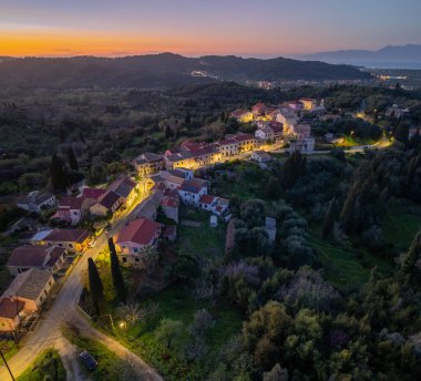 Gökyüzünün alacakaranlık görüntüsü, yemyeşil tepelerin arasında batan güneş tarafından sıcak aydınlatılan binaların bulunduğu tuhaf bir köy yakalıyor..