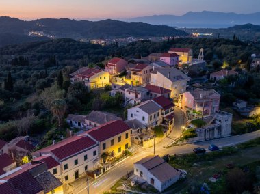 Manzaralı bir köy, tepeler ve sık ağaçların arasına kurulmuş renkli evler ve alacakaranlıkta uzak bir kıyı şeridi ile. Işık sıcak ve davetkar..