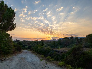 Güneşin turuncu ve sarı ışığın bulutların arasından aktığı ufukta görünen dağlar, yemyeşil araziye doğru bir yol açar..
