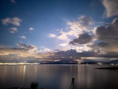 Şehir ışıklarının yansıdığı sakin bir deniz manzarası... Yıldızlarla ve dinamik bulutlarla dolu bir gökyüzünün altında....