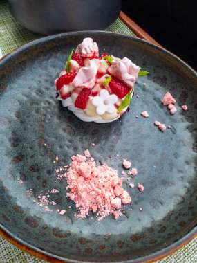 Koyu renkli bir tabakta ustaca hazırlanmış bir tatlı kompozisyonu. Işıklar arasında krema, taze çilek, pembe kırıntı ve çiçek süslemesi yer alıyor.