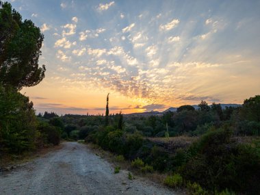 Güneşin turuncu ve sarı ışığın bulutların arasından aktığı ufukta görünen dağlar, yemyeşil araziye doğru bir yol açar..