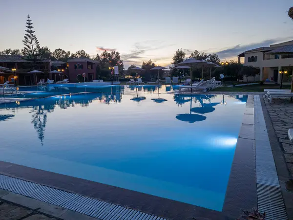 Sakin bir alacakaranlık sahnesinde, çevresindeki binaların, ağaçların ve güneş şemsiyelerinin alacakaranlıktaki yansımaları ile berrak, mavi bir yüzme havuzu vardır..