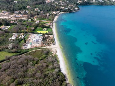 Bu hava manzarası kumlu bir sahile, canlı turkuaz suya, evlere, yemyeşil araziye ve kıyı boyunca bir şantiyeye sahiptir..