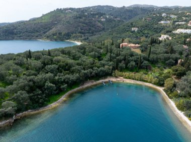 Manzaralı bir hava manzarası, arka planda çarpıcı mavi su, kumlu bir plaj, yoğun yeşil yapraklar ve tepe manzaralı huzurlu bir koy yakalar..