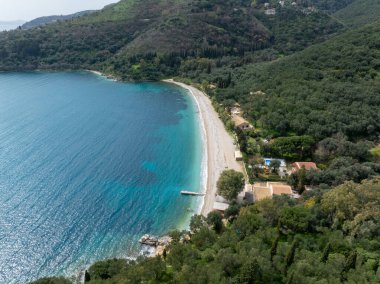 Manzaralı bir hava manzarası kumlu bir plajı, derin mavi denizi ve kıyı şeridi boyunca manzarayı noktalayan yeşil ağaçları gösteriyor..