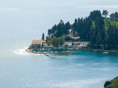 Manzaralı bir yarımadada canlı ağaçlar ve sakin sularla çevrili bir körfeze yerleştirilmiş binalar ve botlar sergileniyor. Huzurlu bir sahil manzarası.