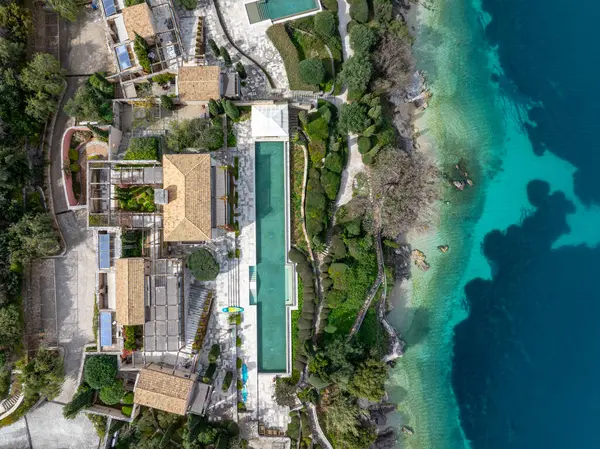 Hava perspektifi, yeşilliklerle çevrili canlı mavi kıyı şeridine bitişik çarpıcı ve uzun bir havuzu olan lüks bir villa yerleşkesini ön plana çıkarıyor..