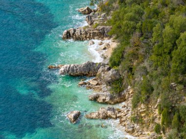 Turkuaz su ve yemyeşil bitki örtüsü olan kayalık bir kıyı şeridinin havadan görünüşü.